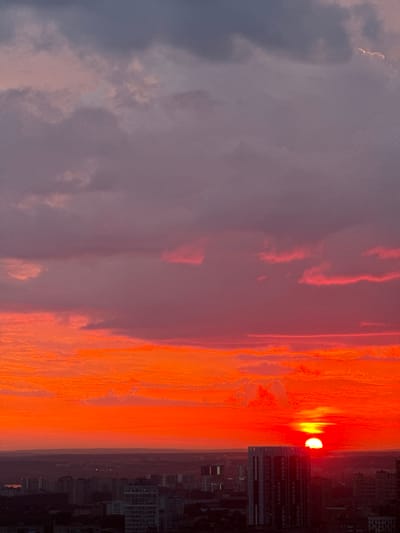 The image depicts a dramatic sunset over a city, likely Savenki, Russia, given the provided context. The sky is ablaze with vibrant hues of orange and red, transitioning to a soft pink and then a deep purple-grey at the top. Wispy clouds are illuminated by the setting sun, creating streaks of light against the darker sky. The sun itself is a brilliant orb, partially obscured by the horizon line, casting a warm glow across the scene.

In the foreground, a silhouette of a city skyline is visible. Numerous buildings of varying heights are discernible, with one taller, modern-looking structure standing out. The cityscape is cast in shadow due to the low angle of the sun. The environment appears to be an urban setting during twilight. There are no visible people or specific actions taking place in the image. The overall impression is one of a natural phenomenon occurring over a populated area.
