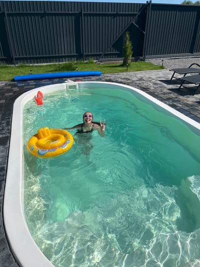 In the city of Sharovechka, Ukraine, a young girl is pictured enjoying a swim in an outdoor pool. She is wearing goggles and a swimsuit, and appears to be in good spirits as she makes a peace sign with her hand. A yellow inflatable ring floats in the pool beside her. The pool is surrounded by a tiled patio and a dark metal fence. In the background, two lounge chairs are visible. The water is a clear turquoise color, and sunlight reflects off its surface.