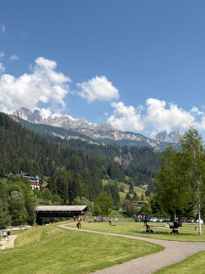 This image captures a serene daytime scene in Soraga di Fassa, Italy, showcasing a picturesque landscape with a tranquil park area. In the foreground, a paved pathway curves through a lush green lawn where several individuals are enjoying the outdoors. One person stands on the path, facing away from the camera with a bag slung over their shoulder. Further ahead, several people are scattered across the grass; some are seated or lying on blankets, a stroller is present, and a young child sits on the grass. To the left, a group of people walks along another path beside a body of water. A covered wooden bridge spans the water in the mid-ground, connecting to the park area. Behind the park, a verdant hillside is dotted with trees and buildings, including a prominent white building with blue-tiled roofs. The background is dominated by imposing, rugged mountains with jagged peaks, their slopes covered in dense evergreen forests. The sky above is a vibrant blue, punctuated by scattered white clouds. The scene conveys a sense of peaceful recreation in a stunning natural setting.
