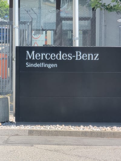 A black rectangular sign with white lettering reads "Mercedes-Benz Sindelfingen". The sign is positioned horizontally, with "Mercedes-Benz" on the top line and "Sindelfingen" on the second line. The sign appears to be made of metal and is mounted on a dark background, possibly a building facade. The sign is set against a backdrop of a building with glass windows and metal fencing. In front of the sign, there is a strip of small white and grey stones. To the left of the sign, a concrete planter or bollard is visible. The ground in front of the planter and stones is paved with asphalt. The overall scene suggests an outdoor setting, likely the entrance to a Mercedes-Benz dealership or facility in Sindelfingen, Germany.