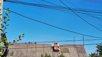 A low-angle, wide shot captures a clear blue sky above a textured, grey corrugated roof in Ialoveni, Moldova. The roof spans the lower half of the image, with a brick chimney, partially covered by a metal patch, rising from its peak. To the left, a green plant with small leaves grows up a pole. Numerous black electrical wires crisscross the sky, creating a complex web against the bright sky. The scene appears to be taken from a street or yard, with some greenery visible at the bottom edge of the frame and on the right side.