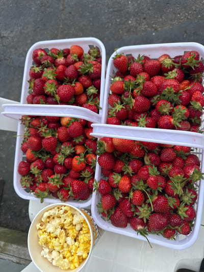 This media file shows two white plastic baskets filled with fresh strawberries. One basket is stacked on top of the other, with the top basket angled towards the viewer. A paper cup filled with popcorn sits to the right of the baskets. The scene appears to be outdoors, with a glimpse of a grey, possibly asphalt surface on the left and a white surface on the right, suggesting a table or counter. The lighting suggests it is daytime. The strawberries are bright red and appear to be ripe, with green stems and leaves visible on some. The popcorn is a light yellow color. No people are visible in the image. The overall impression is one of fresh produce and snacks, possibly at a market or event. There is no visible text in the media file.
