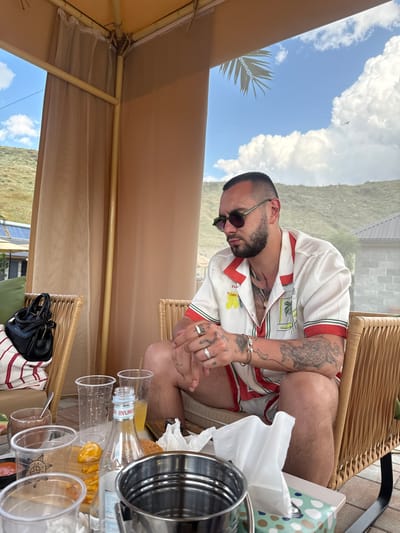 A man is seated at a table outdoors, under a beige canopy with sheer curtains. He wears a white short-sleeved shirt with red and green trim, sunglasses, and several rings. The man has tattoos on his arms and hands. In front of him are several plastic cups, some containing beverages, a bottle of BYUREG sparkling water, and a small amount of food that appears to be fries and a piece of fried chicken. A metallic ice bucket is also on the table. Behind him, a wicker chair is visible. The scene appears to be at a resort or outdoor dining area, possibly near a pool, given the presence of lounge chairs and the open-air setting. The background shows a landscape with hills or mountains and a bright blue sky with scattered clouds, suggesting it is daytime and the weather is clear. The man seems relaxed, looking down at the table.