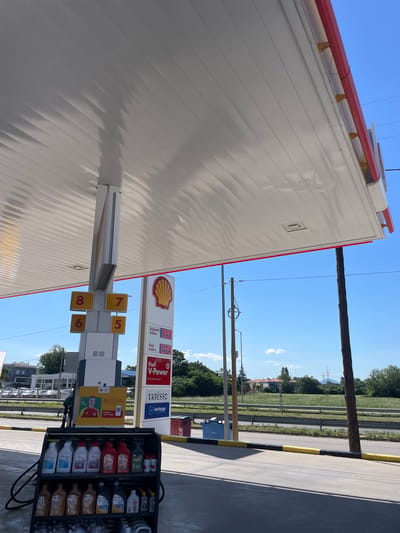 The image depicts a gas station canopy in Komotini, Greece, on a clear, sunny day. The canopy is white with red and yellow trim. Beneath the canopy, a gas pump display shows fuel prices: Unleaded V-Power at 1.899 and Diesel V-Power at 1.549. Adjacent to the pumps, a rack displays various automotive fluids and products, including Shell Helix engine oil and other automotive chemicals. Yellow and black striped barriers line the edge of the paved area. In the background, a grassy field with trees and some buildings can be seen, along with utility poles and overhead wires. The overall scene is static, capturing a typical day at a service station.