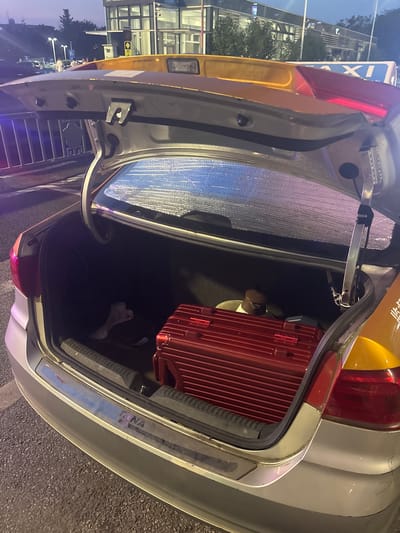 The image shows the open trunk of a silver sedan, which appears to be a taxi based on the yellow and blue signage on the roof. The trunk contains a red hard-shell suitcase, a white plastic bag, and a dark brown thermos or bottle. The interior of the trunk is lined with a reflective material. The car is parked on an asphalt surface, possibly a street or parking lot. In the background, there are buildings, streetlights, and people walking. The overall lighting suggests it is either dusk or evening. Based on the provided information, the location is identified as Xinwu District, China.