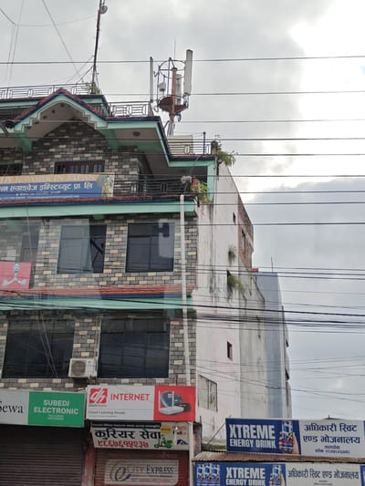 This is a photograph of a multi-story building in Tharpu, Nepal, taken from a low angle, looking upwards. The building appears to be a commercial establishment, with several businesses visible on the lower floors. The building's facade is a mix of brickwork and painted plaster, and it has multiple windows. On the rooftop, there are several communication towers and a network of electrical wires crisscrossing the sky. The sky is overcast with clouds.
On the ground floor, there are storefronts with prominent signage. One sign, in green, reads "SUBEDI ELECTRONIC TALCHOWK 9604198596". Another sign, in red and white, advertises "DishHome INTERNET Cloud Learning House". Below this, there are signs in Nepali script for "कुरियर सेवा" (Courier Service) and a phone number. Further to the right, a sign for "XTREME ENERGY DRINK" is visible, with text in Nepali script that translates to "Official Restaurant and Food Hall". This sign also has a phone number. Another similar sign for "XTREME ENERGY DRINK" is located below the first one.
On an upper floor, a banner reads "STUDY & WORK IN JAPAN" in English and Nepali. There is also a smaller sign with a phone number and email address. A framed image of a person in traditional attire is also visible on this level. Further up the building, there are more windows and what appear to be air conditioning units. The overall impression is of a busy commercial area within a town.