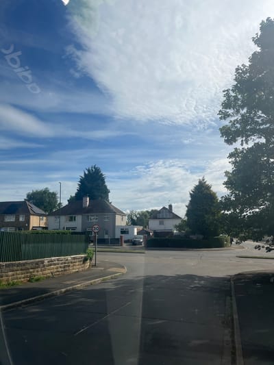 This image captures a residential street scene in Nottingham, United Kingdom, under bright daylight. The view is looking down a street with houses on the right side and a line of trees on the left. The sky, filled with white, wispy clouds (likely cirrus and stratocumulus) against a blue background, occupies a significant portion of the upper frame.
On the right side of the road, several multi-story houses are visible, featuring pitched roofs and varying architectural styles. Some houses have visible windows, and at least one appears to have solar panels on its roof. A green slatted fence runs along the immediate right foreground, behind which a white car is partially visible. Further down the street, another silver or grey car is parked.
The asphalt road, which appears to curve to the right in the distance, takes up the lower portion of the image. It has a white line marking the edge and a pavement or sidewalk running alongside it. Shadows from unseen objects or structures are cast across the road. A stone retaining wall or terracing is visible between the green fence and the pavement.
A prominent red circular road sign with "30" in white, indicating a 30 mph speed limit, stands near the houses on the right. Tall trees with dark green foliage frame the top and left side of the image, and additional trees are scattered among the houses.
No people or specific activities are visible in the scene, which appears quiet and residential. The weather is sunny with scattered clouds, indicating a clear day, likely late morning or early afternoon given the quality of light and shadows. A faint, reversed reflection of what appears to be "obibp" (possibly "Adidas") is visible in the bottom left corner, suggesting the photo was taken through a window.