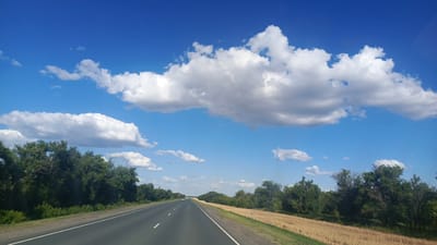 This media file depicts a scenic view of a road in Золотая Степь, Russia, under a bright blue sky dotted with fluffy white clouds. The scene appears to be a daytime shot, likely late morning or early afternoon, given the clear visibility and the intensity of the sunlight.
The primary subject is a paved road, extending into the distance and curving slightly to the right. The road has clear white lane markings, indicating it's a well-maintained route, possibly a highway or a major rural road. To the left of the road, dense green trees form a lush border, with some foliage extending over the road's edge. To the right of the road, a field of dry, golden vegetation, likely harvested crops like wheat or barley, stretches out, contrasting with the greenery on the other side. The horizon is lined with more trees and vegetation, suggesting a rural or semi-rural landscape.
The sky is a dominant feature, with large, cumulus clouds scattered across it. These clouds are voluminous and well-defined, with some casting soft shadows. The overall mood of the image is peaceful and serene, evoking a sense of journey and openness. There are no people or animals visible in the frame, and no objects beyond the natural landscape and the road. The setting is clearly outdoors, in a natural environment. There is no visible text in the image.