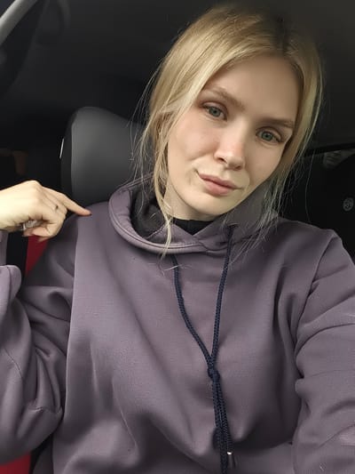 The image shows a young woman with blonde hair, blue eyes, and light skin, wearing a purple hoodie. She is in the driver's seat of a car, as indicated by the car interior in the background. Her left hand is raised, and she is pointing towards her left shoulder with her index finger, wearing rings on her fingers. The woman is looking directly at the camera, with a neutral expression. The background of the image is blurred, but it appears to be the interior of a vehicle. There are no other people or objects clearly visible in the background. The lighting in the image suggests it was taken during the daytime, possibly with natural light coming from outside the car. The overall setting is a car interior in Ягул, Russia.