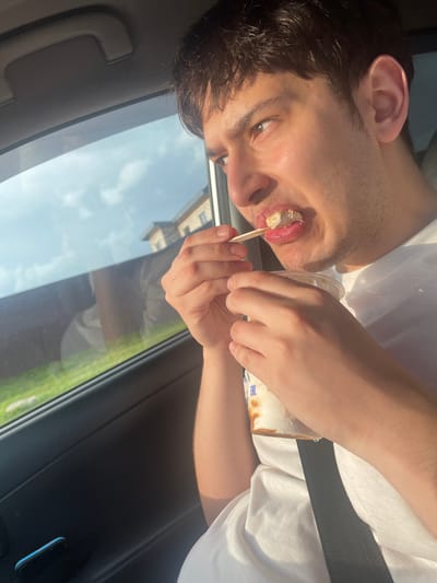 A young man is shown from the chest up, inside a vehicle, looking out the passenger-side window. He is holding a small wooden spoon with food on it and bringing it to his mouth. He is wearing a white t-shirt. Outside the window, green grass and a building are visible, suggesting a suburban or rural setting, potentially in Gavrilovka, Russia. The interior of the car is dark, with the window reflecting the sky.
