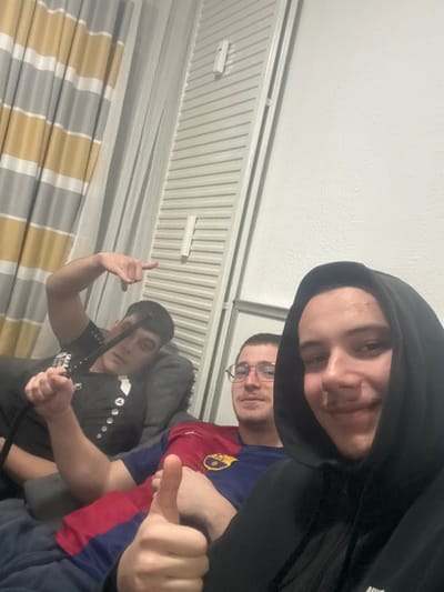 This indoor photograph captures three young males in a casual setting in Krems an der Donau, Austria.
In the foreground on the right, a male wearing a black hooded sweatshirt with a visible Nike logo smiles at the camera and extends a right thumb in an upward gesture. Immediately to his left, a second male, wearing glasses and a blue and red FC Barcelona jersey, looks towards the camera with a subtle smile. To the left and slightly behind the second individual, a third male, dressed in a black shirt with white button-like embellishments, holds a black hookah hose in his raised left hand. His right arm is also raised, with the index finger extended, pointing to the left. A faint vapor plume is discernible around the hookah hose.
The background consists of a light-colored, textured wall on the right. To the left of the wall, a vertical white slatted structure, possibly a bifold door or interior shutter, is visible, featuring two vertically mounted white rectangular objects, potentially sensors or fixtures. Further left, partially drawn striped curtains in yellow/gold and grey hues are visible. The individuals appear to be seated on dark-colored upholstered furniture. The scene is illuminated by interior lighting.