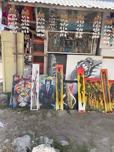 An outdoor art stall displays various paintings. Subject matter includes wildlife, such as lions, zebras, and giraffes, alongside human figures. Some pieces feature abstract or surreal elements, like a gorilla in a suit or a skull merging with a guitar. The paintings are displayed on the ground and leaning against structures, suggesting an informal sales setting. Decorative shell ornaments hang above the artwork.