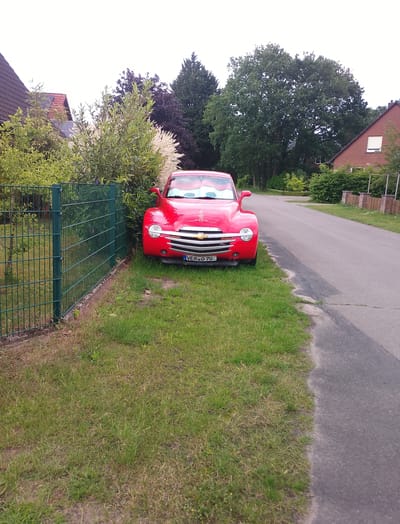 The image depicts a red Chevrolet SSR pickup truck parked on a grassy area next to a paved road. The truck is angled towards the road, with its front end visible. A green metal fence runs along the edge of the grassy area. In the background, there are trees, bushes, and houses, suggesting a suburban or rural setting. The license plate of the truck reads "VER O 70". The sky is overcast. The image was taken in Langwedel, Germany.