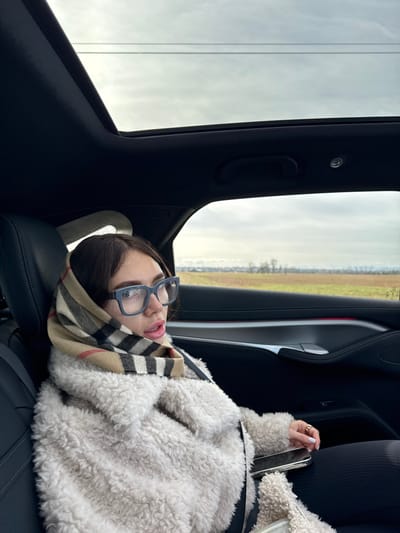 A female individual is seated in the rear passenger compartment of a vehicle, with the presumed location being Saint-Bonnet-de-Mure, France. The female, with dark hair partially covered, wears light blue-framed eyeglasses and a beige, black, and red plaid pattern scarf draped over her head and neck. She is dressed in an off-white, textured, possibly faux fur, jacket over dark trousers. A seatbelt is secured across her chest and lap. A gold-colored ring is visible on her right ring finger, and her fingernails are painted a light color. A black smartphone rests on her lap. The vehicle's interior features dark upholstery, a panoramic sunroof, and a dark door panel with a silver-colored trim and a faint red accent light. Outside the window, an overcast sky with power lines is visible above a flat landscape consisting of brown agricultural fields and distant bare trees. The individual is looking towards the left side of the frame with her mouth slightly parted.