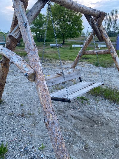The image shows a rustic wooden swing set constructed from natural logs. The swing seat is made of weathered wooden planks suspended by metal chains. The ground beneath the swing is composed of gravel and dirt, with sparse green vegetation growing in patches. In the background, there are trees, a grassy area with some benches, and a cloudy sky. The location is identified as Chkalovka, Armenia. There are no people visible in the image. The scene appears to be an outdoor recreational area, likely a park or a yard.