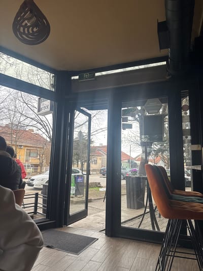 The image depicts an interior view of a commercial establishment, likely a cafe or bar, in Kumanovo, North Macedonia, looking out through a glass facade towards a street scene on an overcast daytime.

In the foreground, partial views of at least two individuals are visible with their backs to the camera on the left. One person wears a dark jacket over a red garment, while another wears a light-colored top. On the right, two high-backed bar stools with black metal frames are present; the closest is upholstered in orange fabric, and the one behind it in dark green. The interior floor is covered with light brown, wood-patterned tiles, with a dark grey welcome mat positioned at the threshold of the entrance. The ceiling is off-white, featuring a dark, open-lattice, teardrop-shaped light fixture in the upper left and a black, exposed circular ventilation duct along the upper right. A green "EXIT" sign with a running figure pictogram is affixed above the central glass door.

The exterior scene, visible through the dark-framed glass doors and windows, shows a paved street or sidewalk. Deciduous trees with bare branches line the street, indicating late winter or early spring. Across the street, residential buildings with red tiled roofs are visible, including a prominent two-story yellow building with a balcony. A white sedan is parked or moving on the street. Outside the establishment's entrance, a dark cylindrical planter containing purple flowers is situated. Various decals and reflections are visible on the glass, including a "ZIPP SPOT" logo, a grey hexagonal sign, and an advertisement featuring a green beer bottle (identifiable as "SKOPJESKO"). A vertical rectangular sign with Cyrillic text, partially readable as "SMOOTH," is also visible through the left glass panel. Some loose bricks or debris are scattered on the ground near the right side of the exterior view.