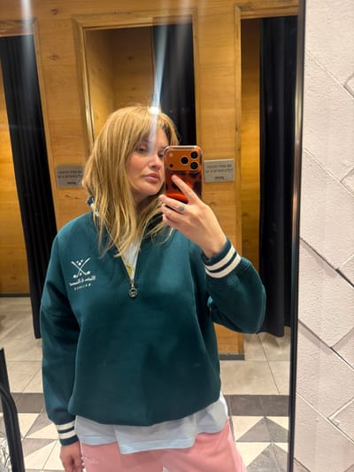 A female individual is depicted taking a mirror selfie inside what appears to be a changing room or retail establishment in Nahariya, Israel. The individual has light brown, shoulder-length hair with bangs, and is looking directly towards the camera reflected in the mirror. She is wearing a dark teal quarter-zip sweatshirt featuring a white embroidered logo of two crossed golf clubs and inverted Hebrew text (possibly "BALAMCE" or similar, reading "BALANCE" when upright) on the left chest. The sweatshirt has white and dark teal striped cuffs. A light blue t-shirt is visible beneath the sweatshirt, and pink sweatpants are partially visible at the bottom of the frame. The individual wears a ring on her left hand's ring finger and holds a smartphone with a tortoiseshell-patterned case and a triple-lens camera system, used to capture the image.

The reflected environment shows wooden paneled walls on the left, with dark fabric curtains or partitions visible through openings, suggesting changing stalls. On the right, a textured wall with a light-colored chevron or zig-zag pattern is visible. The floor is tiled with a geometric pattern, likely black and white or grey. Hebrew text signs are visible on the wooden paneling, stating "נא לא להכניס מזון ושתיה" (Please do not bring food and drink) and "נא לא לעשן" (Please do not smoke), confirming the Israeli location. A distinct vertical light flare is visible in the reflection above the individual's head, originating from an overhead light source.
