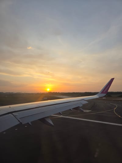 The image shows an airplane wing with the "wizzair.com" logo, presumably taken from inside the aircraft. The plane is on the runway, and the sun is setting in the background, with its light reflecting on the wing. The runway is visible below the wing. The image was not taken in North Hertfordshire, United Kingdom because it contains a runway.