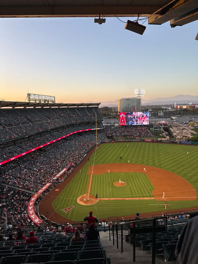 The image captures a view of Angel Stadium in Anaheim, California, during a baseball game at sunset. The stadium is filled with spectators seated in tiered rows of dark seats. Red banners with the "Angels" logo are visible along the stadium's perimeter. The baseball field is illuminated, with players on the pitcher's mound and other positions. A large video screen displays images of people. In the background, a cityscape with buildings and mountains is visible under a soft orange and blue sky. The foreground shows empty stadium seating and a railing.