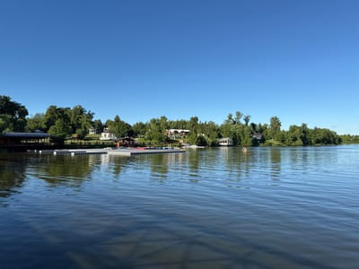 The image shows a wide, calm body of water with ripples on its surface, reflecting the blue sky and lush green trees lining the opposite bank. In the foreground, the water is a deep blue. Along the shore, there is a long, modular floating dock made of grey plastic sections. Several buildings are visible nestled among the trees on the far bank. To the left, a wooden structure with a dark roof extends over the water, appearing to be a boathouse or pier. Further along the shore are white gazebos and other buildings, suggesting a resort or recreational area. In the middle of the water, a small orange and red boat with at least two people is visible, paddling away from the shore. The sky is a clear, bright blue with no clouds. The scene depicts a tranquil summer day at a waterfront location, possibly in Grebnevo, Russia, given the presence of what appears to be a recreational facility.