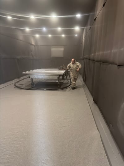 A person wearing a camouflage protective suit is standing in what appears to be a spray-painting booth. The person is bald and appears to be holding a spray gun, directed towards a long, rectangular object supported by a metal frame with wheels. This object seems to be a large panel or piece of furniture, painted white with several horizontal slats. The interior of the booth is dimly lit by several rectangular lights positioned along the ceiling. The walls and ceiling of the booth are dark gray, and the floor is a lighter, speckled gray. The overall impression is a professional, controlled environment for industrial painting. The image is taken vertically, with the person and the object appearing in the center, and the booth extending into the background. There is no visible text in the image.