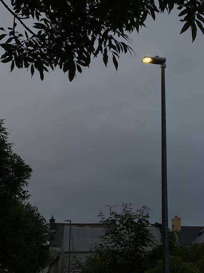 A street lamp with a yellow light illuminates a dull, overcast sky. In the foreground, dark silhouettes of trees frame the scene, their leaves dense and detailed against the muted sky. To the right, the corrugated metal roof of a building is visible, along with parts of its weathered wall. The building appears to be older, with patches of peeling paint and exposed brickwork. Further to the right, more trees and foliage are present, also in silhouette. The overall impression is one of twilight or a very gloomy day. The lighting suggests it is either dusk or dawn, or a particularly dark, cloudy afternoon. The presence of the street lamp indicates an outdoor, likely urban or suburban, setting. No people are visible, and the scene is static, with no specific activity taking place. The location context of Mountcharles, Ireland, suggests a temperate climate, consistent with the overcast sky.