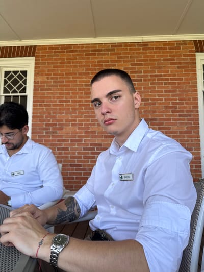 Two individuals, identified by nametags as "MIKE" and "SIMEON", are seated outdoors on a porch, with a brick wall and a white-trimmed window as the backdrop. Both are wearing white button-down shirts. Mike, on the left, is wearing glasses and looking down. Simeon, on the right and closer to the camera, is looking forward. He has a buzz cut and visible tattoos on his left arm, and is wearing a watch and a red string bracelet. The scene appears to be in Harbor Springs, Michigan, United States.