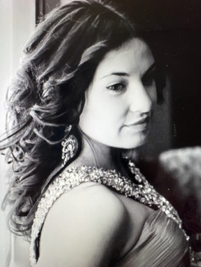 This black and white portrait features an adult woman, shown in what appears to be a formal setting. The image is oriented vertically, with the woman's head tilted towards the right side of the frame. She has medium-length, dark, wavy hair that cascades around her face and shoulders. Her expression is composed, with a subtle smile or smirk, and she appears to be looking slightly away from the camera.
She is dressed in an elegant garment, possibly a formal dress or top, which is adorned with intricate, sparkling embellishments along the neckline and shoulder strap. These details are a prominent feature of her attire. The lighting strongly illuminates her face and the decorated portion of her clothing, creating a contrast with the dark, blurred background. This selective focus emphasizes the subject. The image displays a fine grainy texture, characteristic of certain photographic processes or scans.
The scene captures a posed moment, suggesting a photoshoot or a special event. There are no other discernible objects or visible text within the frame. The overall mood conveyed is one of elegance and serenity. The provided location context for this media file is Родниковское сельское поселение, Ukraine.