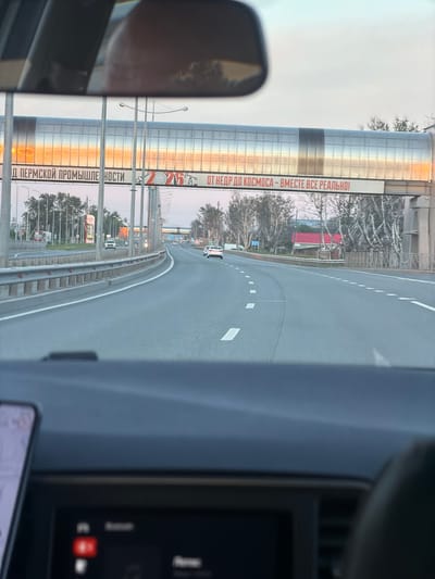 The image captures a view from inside a vehicle driving on a highway in Crokhovo, Russia. A large pedestrian bridge spans the highway, decorated with the inscription "ОТ НЕДР ДО КОСМОСА - ВМЕСТЕ ВСЕ РЕАЛЬНО!" and "Д ПЕРМСКОЙ ПРОМЫШЛЕНОСТИ". Several other vehicles are on the road. The environment includes trees alongside the road and urban buildings in the distance. A car's side mirror and dashboard details are visible in the foreground.