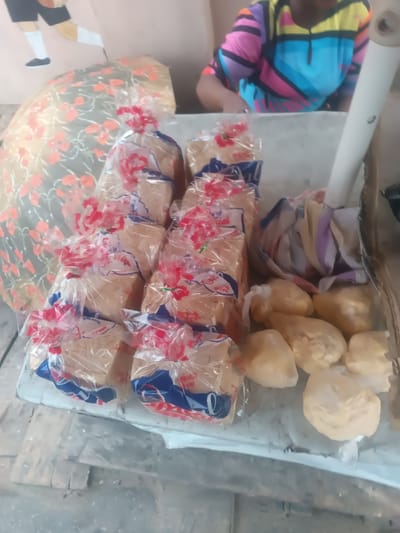 A table laden with loaves of bread in clear wrappers, each adorned with red and white ribbon. Alongside the bread, a pile of soft, golden-yellow dough sits wrapped in plastic. The table appears to be in a brightly lit area, with glimpses of a colorful painting in the background. A person wearing a multicolored shirt can be seen partially in the upper right corner.