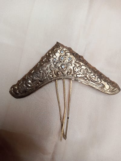 A gold-toned metallic hair ornament, likely a comb or tiara fragment, is centrally depicted against a light beige, subtly textured fabric background. The ornament features an ornate, roughly triangular or crescent-shaped body, approximately 15-20 cm in width, embossed with intricate floral and scroll patterns. Seven small, light-blue or clear rhinestones are set within a prominent central floral motif. Three thin, flat, gold-colored metal prongs extend downwards from the ornament's base, forming a comb structure; one prong exhibits a slight bend and discoloration. The metal shows signs of tarnish and wear. The object is static, with no visible individuals or active interactions. The environment is an unidentifiable indoor setting within Qarabucaq qəsəbə, Azerbaijan, indicated by the provided context.
