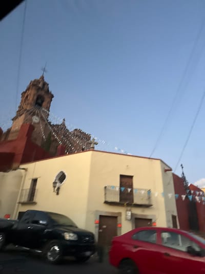 An upward, angled view shows a light-colored building with a red roof, adorned with hanging decorations. Adjacent is a church steeple against a pale blue sky. A red car is partially visible on the right, alongside a black car parked in front of the building. The perspective gives a distorted impression of the scene.