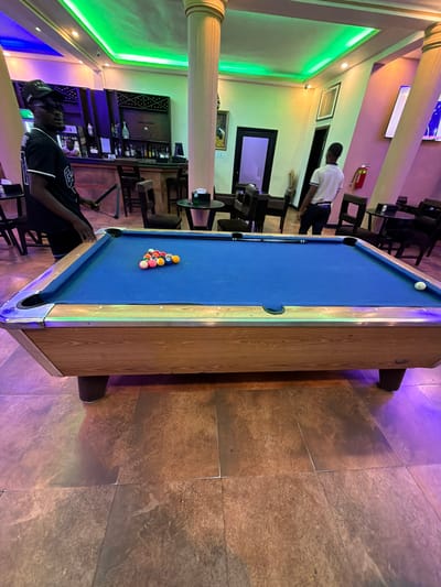 The image shows a pool table in what appears to be a bar or lounge. The table is set up with a rack of balls, ready for a game. A man is sitting at one end of the table, holding a pool cue, seemingly preparing to play. Another man is walking in the background near a fire extinguisher. The background also shows a bar area with bottles and seating. The floor has a tile pattern. The location is suspected to be in Monrovia, Liberia.