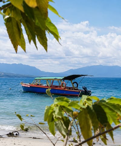 A colorful boat with a green canopy is anchored in the clear blue waters of Lumban Bul Bul, Indonesia. The boat has a red hull with a blue stripe and a yellow frame supporting the canopy. A life preserver is visible on the side of the boat. In the background, hazy blue mountains rise against a sky filled with white, fluffy clouds. In the foreground, lush green leaves and branches of a tree frame the scene, with the sandy shore and gentle waves of the lake visible at the bottom. A black outboard motor is attached to the stern of the boat.