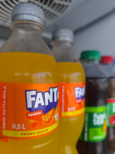 The image shows several bottles of beverages lined up inside a refrigerator. The focus is on the front bottle, which is a 0.5 L bottle of Fanta Orange. The label on the Fanta bottle is primarily orange and purple and features the Fanta logo in white letters. Below the logo, it says "Apelsin" (which means orange in Azerbaijani) and "PROMO AKSIYA" (which means "Promo Offer" in Azerbaijani). The liquid inside the Fanta bottle is a bright orange color.

Behind the Fanta bottle, other beverage bottles are visible, including a bottle with a green cap that appears to be a Fuse Tea. The background is the interior of the refrigerator, with a ribbed, white surface at the top, suggesting shelves or cooling elements. The overall scene is brightly lit, indicating the refrigerator light is on, and the time of day is not discernible. The setting is likely a store or a home refrigerator. There are no people visible in the image. The image is captured from a low angle, looking up at the bottles.