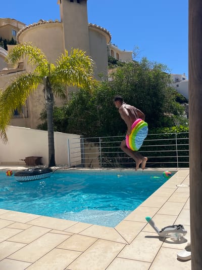 A man is jumping into a swimming pool while holding a stack of inflatable swimming rings. The pool is surrounded by light-colored tile. A snorkel and flippers are on the deck of the pool. A palm tree is near the pool, and there is a house in the background. The sky is blue and sunny. A tree, a beach ball, and a black rubber tire float in the pool. The location is el Poble Nou de Benitatxell / Benitachell, Spain.