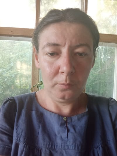 The media file is a close-up selfie of a middle-aged woman. She has dark hair pulled back and is looking down with a neutral expression. Her skin appears somewhat flushed, and there are visible lines on her forehead. She is wearing a dark blue dress with a round neckline and a small button closure. The dress also has a patch pocket on the left side of the chest. The background suggests an indoor setting with a window behind her. Through the window, lush green foliage and trees are visible, indicating an outdoor scene with daylight. The lighting in the image suggests it was taken during the daytime, possibly late morning or afternoon, with natural light filtering in from the window. There is no visible text in the image. The overall impression is a candid, unposed portrait taken in a home or a similar casual environment. The location context provided is Berezivka, Ukraine.