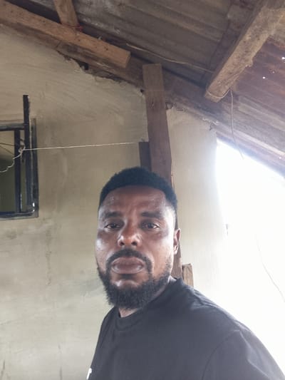 A medium shot captures a Black man with short, dark hair and a full beard, looking directly at the camera. He is wearing a plain black t-shirt. The background consists of a textured, light-colored wall, a small window with a dark frame, and overhead a corrugated metal roof supported by wooden beams. The lighting suggests it is daytime, possibly with bright sunlight coming from the right, creating some overexposure in that area. The overall scene appears to be outdoors or in an open-air structure, possibly under construction or in a rustic setting in Adikpo, Nigeria. There is no discernible text in the image.