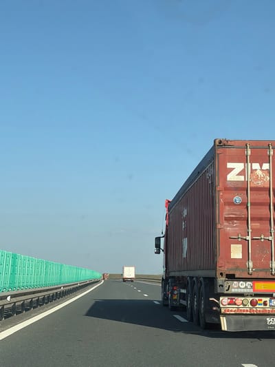 The scene depicts a multi-lane asphalt highway near Borcea, Romania, observed under a clear blue sky. A red semi-trailer truck, bearing a "ZIM" logo on its weathered cargo container, is prominently positioned on the right side of the frame, proceeding away from the viewer. The truck's rear displays multiple red and amber lights, reflective striping, a partial "SCANIA" logo on its mudflap, and a sticker featuring speed limits (50, 70, 80 km/h) and the flag of Romania. To the left, a continuous green modular sound barrier runs parallel to the highway, situated above a metal guardrail. Further down the highway, a white semi-truck is visible in the distance, also traveling in the same direction. The roadway includes distinct white lane markings. No individuals are present in the visible area.