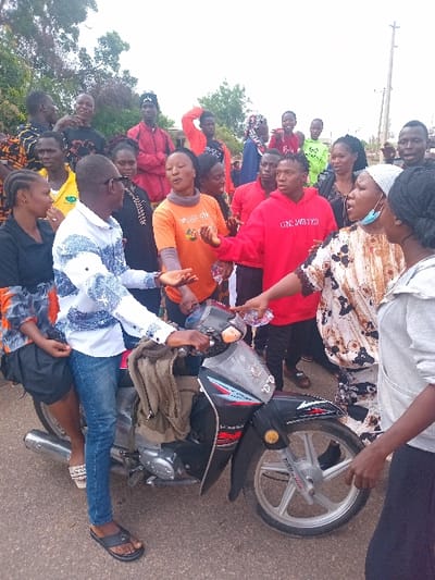 The scene is in Ungwan Tanko, Nigeria, at the intersection of a paved road and a dirt path. A group of about 15 young adults are gathered around a black motorcycle. The motorcycle is facing left and has a large black bag attached to the back. A young man in a white shirt with blue jeans is standing on the left side of the motorcycle, holding the handlebars. A woman in a blue and white patterned blouse is standing on the right side of the motorcycle, seemingly interacting with another woman wearing a grey hooded sweatshirt. Many other people are standing around, some engaging in conversation, others looking toward the camera. Some people are wearing traditional Nigerian clothing, while others are wearing more modern clothing. The scene is brightly lit, and the weather appears to be pleasant. A utility pole stands behind the group, and a few trees and other vegetation can be seen in the background. The individuals appear to be casually interacting with one another.