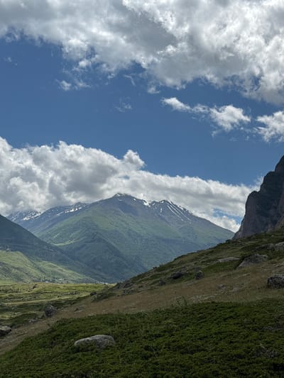 This vertical shot captures a vast mountain landscape on a cloudy day, possibly near El'tiubiu, Russia. The sky is a vibrant blue, dotted with fluffy white clouds. The foreground is dominated by a rocky, grassy slope covered in low-lying green shrubs and scattered boulders. As the eye moves towards the background, the terrain transforms into rolling green mountains, some with patches of snow on their peaks. The distant valley reveals a cluster of small houses, suggesting a settlement. The light suggests it is daytime, likely afternoon given the contrast of light and shadow.