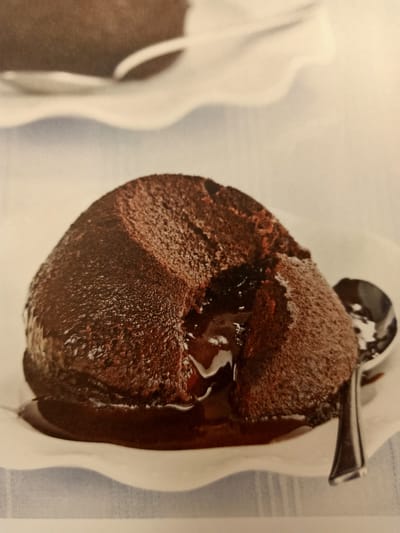 The image displays a close-up shot of a single chocolate lava cake on a white dessert plate. The cake has a rich, dark brown exterior, dusted with a lighter brown powder, likely cocoa powder or powdered sugar. The top of the cake has a slight crack, revealing a molten, dark chocolate interior that is oozing out onto the plate. A silver spoon rests on the right side of the plate, partially submerged in the chocolate sauce. The plate itself has a ruffled edge and sits on a surface covered with a light blue and white striped cloth. The background is a soft, blurred off-white. There are no people visible in the image. The scene is set indoors, with soft lighting. There is no information or visual cues within the image to suggest a location in Rumaithiya, Kuwait.