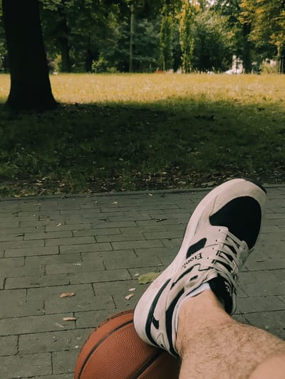The image shows a close-up perspective of a person's leg, wearing a white and black athletic shoe, resting on a basketball. The shoe has the brand name "Eton" visible on its side. The basketball is orange and textured. The leg is covered in light brown hair. The ground beneath is paved with grey rectangular bricks, with a few fallen leaves scattered on them. In the background, a grassy area transitions into a park with trees and dappled sunlight, suggesting an outdoor urban setting, possibly in Sosnowiec, Poland.