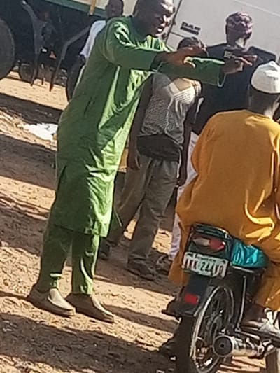 In a daytime scene, likely in Gadao, Nigeria, several individuals are gathered outdoors. The foreground features a person wearing a bright yellow top and blue trousers, seated on a motorcycle. The motorcycle has a license plate with the text "AKK 247V9". Another person, dressed in a long green outfit, stands to the left, gesturing with their hands. This individual appears to be the central figure of focus. Behind them, other people are visible, some in traditional attire. The ground is covered in sandy earth, suggesting a dry and possibly dusty environment. The lighting indicates it is daytime. The overall mood seems to be one of interaction or a gathering, with the prominent gesture of the person in green suggesting a conversation or direction.
