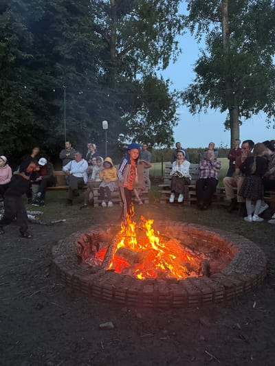 A group of people are gathered around a bonfire in a rural outdoor setting, possibly in Завалочыцкі сельскі Савет, Belarus, during dusk. The bonfire is in a circular brick fire pit in the foreground, with visible flames and burning logs. Several individuals are seated on wooden benches made of pallets, and others are standing. In the left foreground, a young person in dark clothing is captured mid-motion, possibly dancing or interacting with the fire. Another young person wearing a blue baseball cap stands near the fire, facing the camera. The background features trees and a dusky sky. The ground appears to be dirt and grass. String lights are visible hanging from trees in the upper portion of the image.