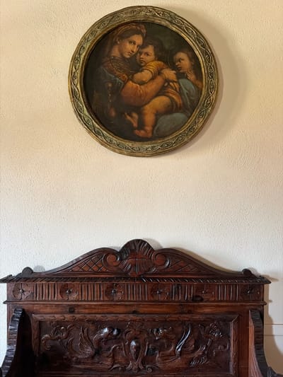The image displays a circular painting of a Madonna and Child, framed with an ornate, aged gold-colored border. The painting itself depicts a mother cradling two children, rendered in warm, earthy tones with a somber, emotional quality. The mother's gaze is directed outward, conveying a sense of tenderness and perhaps melancholy. The children are nestled close, their faces soft and serene. The painting is hung on a textured, off-white wall, with a soft shadow cast to its left, suggesting ambient lighting.

To the right of the painting, a large, intricately carved wooden object is visible. It appears to be a piece of ornate furniture or decor, possibly a cabinet or a decorative panel. The wood is dark and rich, with deep carvings depicting floral motifs and possibly a mythical creature or dramatic scene on the rightmost section. The carvings are detailed and three-dimensional, showcasing skilled craftsmanship. The object has a stepped or shelf-like element at the bottom, further suggesting it might be a piece of furniture.

The scene is indoors, likely in a room with a neutral color palette, given the prominent white wall. The lighting appears to be soft and diffused, creating subtle shadows that highlight the textures of both the wall and the wooden carving. The overall impression is one of quiet elegance, with a focus on classical art and detailed artisanal work. The image is static, capturing a moment in time with no active events taking place. No visible text is present in the image. The location context provided is Asciano, Italy, which might suggest the style of art and craftsmanship seen in the image.