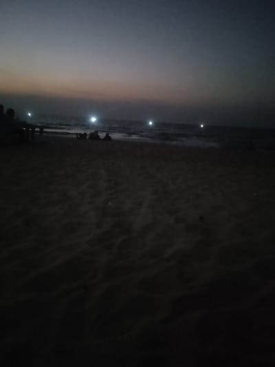 This image captures a nocturnal beach scene in Al-Mawasi, Palestinian Territories. The upper portion of the frame displays a dark twilight sky, transitioning from a deep grey-blue at its zenith to a lighter, reddish-grey hue at the horizon. The middle ground features the sea, exhibiting a relatively calm surface with gentle waves breaking near the shoreline. Four distinct, bright white light sources are visible across the water, appearing as illuminated points. The foreground and lower half of the image consist of dark, textured sand. On the far left, two human silhouettes are vaguely discernible, positioned near a dark, table-like object. Further to the right, closer to the water's edge, a group of three to four additional human silhouettes are observed, appearing to be seated or reclining on the sand, facing the sea. No specific actions or interactions are clearly identifiable due to the low light conditions.
