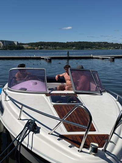 The image shows a white speedboat docked at a pier on a sunny day. Three shirtless men are seated in the boat. One man is at the helm, another is seated in the passenger area, and a third man is visible from the chest up behind the windshield. The boat has a purple cover on the dashboard and water droplets are visible on the windshield. In the background, there is a body of water with several floating docks and a distant treelined shore with buildings. The sky is blue with a few scattered clouds. This scene is likely located in Tønsberg, Norway, given the presence of a harbor and boats.