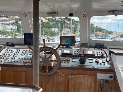 The image shows the interior of a boat's bridge, likely in the city of Le Lavandou, France, overlooking a harbor filled with sailboats. The bridge features a wooden steering wheel, a navigation screen, various control panels with switches and buttons, a radio, and a microphone. The background reveals buildings on a hillside and more boats in the water. A "no smoking" sign is visible on the left side of the image. The overall scene is that of a functional maritime control center.