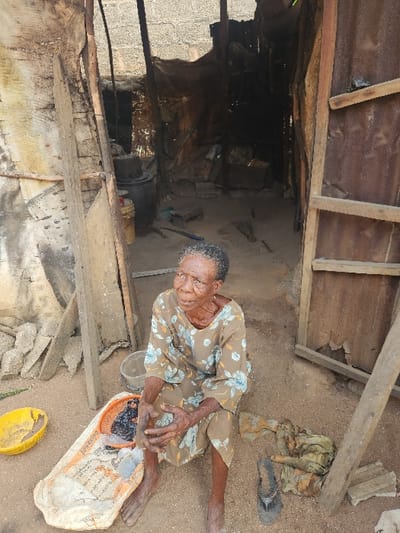 The image depicts an elderly woman sitting on sandy ground in a humble, semi-outdoor setting in Umuoye, Nigeria. The woman, appearing frail with wrinkled skin, wears a patterned dress with light blue floral designs on a brown or beige background. She looks towards the viewer with a calm, neutral expression, and her hands are clasped or resting on her lap.
In front of her right leg is a large, light-colored tray or platter, which appears to be a repurposed bag. It contains a dark, granular substance, and faint text on it includes "CEMENT" and possibly "SUPER." Nearby, a yellow bowl or lid rests on the ground. Behind her, a single dark sandal and a piece of dirty, olive-green fabric are visible on the sand.
The setting consists of a makeshift structure with wooden planks and beams forming supports and part of a roof or shelter. Corrugated metal sheets are also visible as part of the construction. The ground is sandy or dirt, with scattered concrete blocks and debris. In the background, there is a dark, unlit interior space, possibly a room or storage area, further supported by rough wooden elements. The scene is lit by natural daylight, suggesting it is daytime. The overall environment conveys a sense of basic or impoverished living conditions.